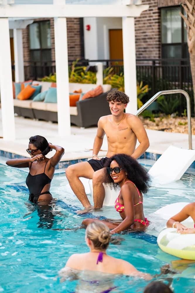 A group of friends enjoying a pool, with one man sitting on the edge and three women in the water, smiling and wearing swimsuits.
